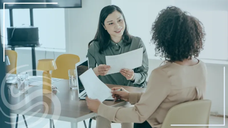 Two female colleagues setting at a conference room table with reports, a laptop, glasses of water, and a lamp in an office setting with the Specialized Recruiting Group Logo